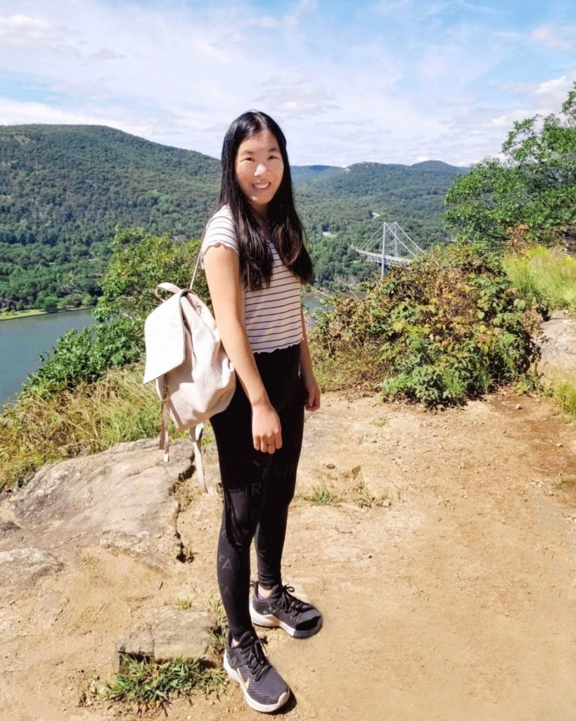 A girl stands on top of a mountain. She is wearing a black and white striped shirt, black leggings, sneakers, and a blush pink backpack.