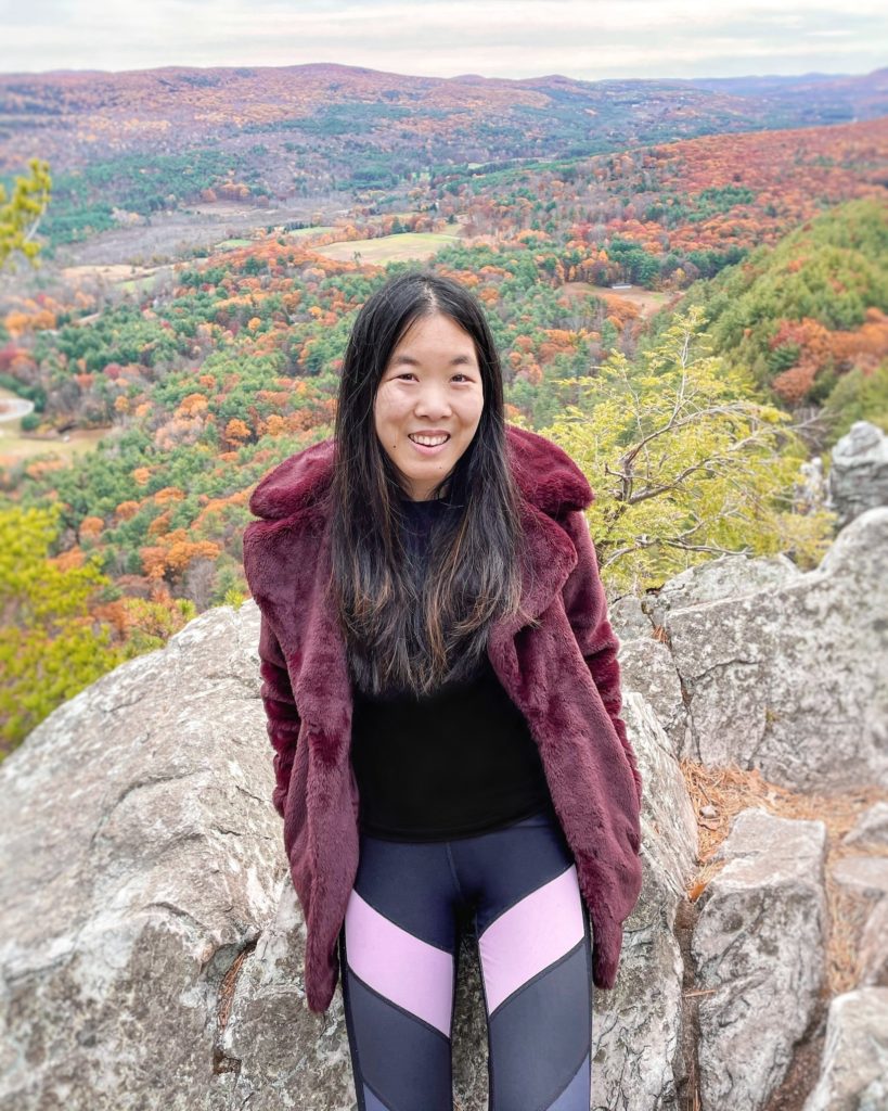 A girl sits on a rock on top of a mountain, overlooking fall colors of orange, yellow, and green. She is wearing a burgundy faux fur coat, a black shirt, and gray and pink colorblock leggings.