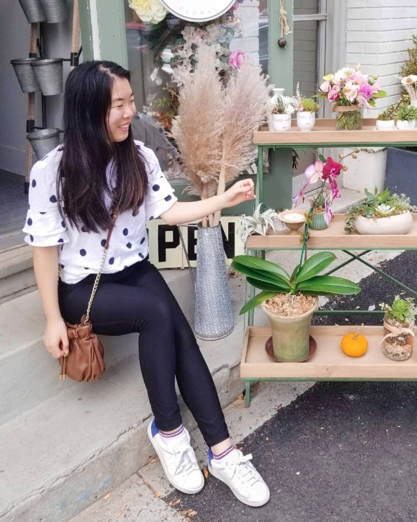 A girl sits on a stoop. She is wearing a black and white polka dot shirt, black leggings, and white sneakers.