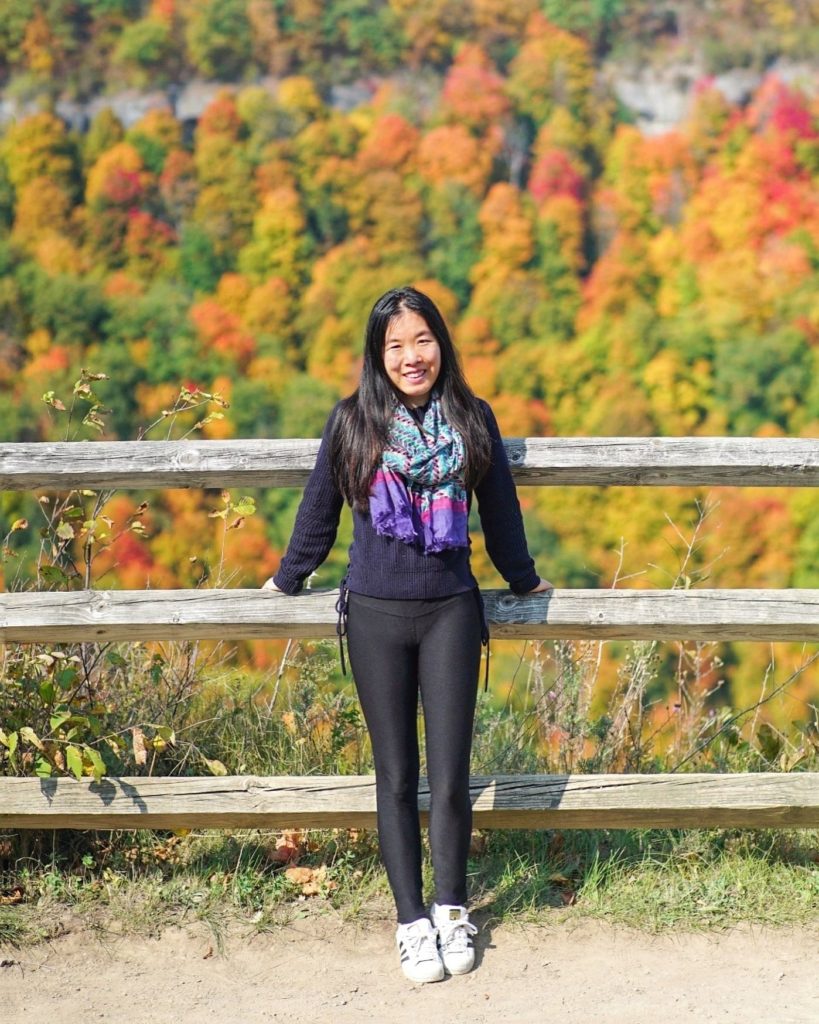 A girl stands along a fence overlooking peak fall foliage colors of red, orange, and yellow. She is wearing a navy sweater, black leggings, white sneakers, and a blue floral scarf.