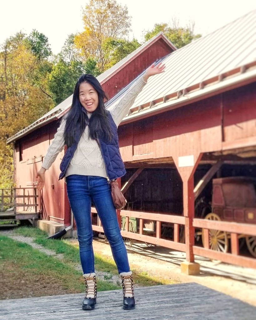 A girl poses in front of a red barn. She is wearing a cream cable knit sweater, navy vest, blue jeans, and black ankle boots.