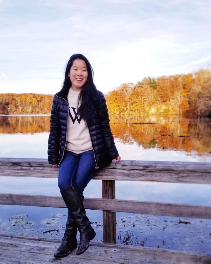 A girl sitting on a bench in front of a lake. She is wearing a black puffer coat, white sweater, jeans, and boots.